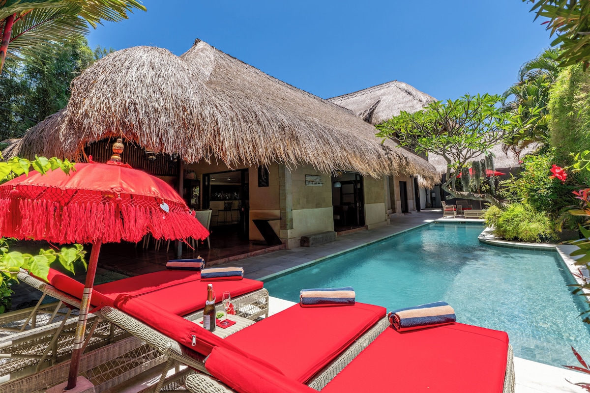 A poolside area with red lounge chairs, a thatched-roof structure, and a clear blue pool.