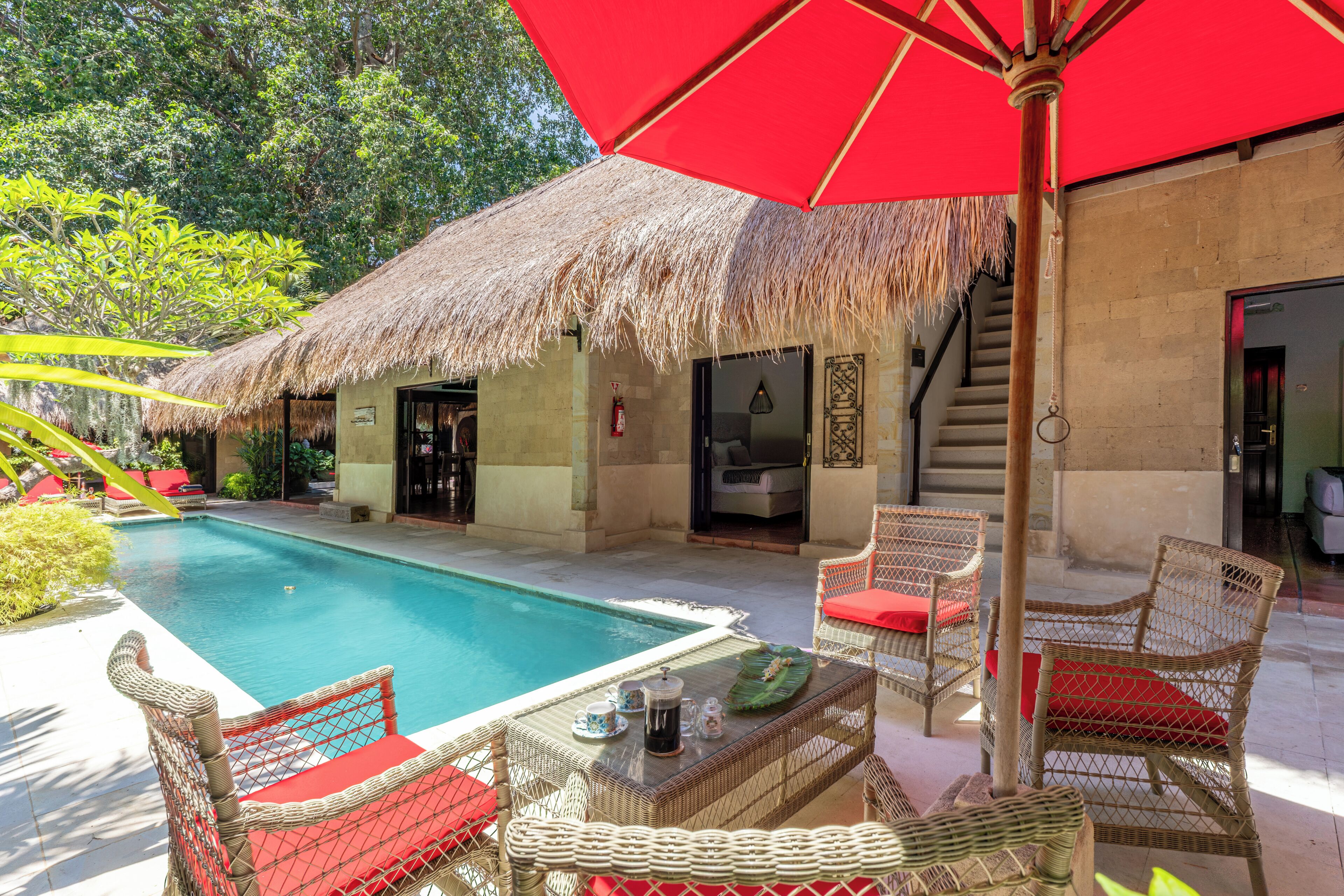 A poolside area with a thatched-roof structure, wicker furniture, and a red umbrella.