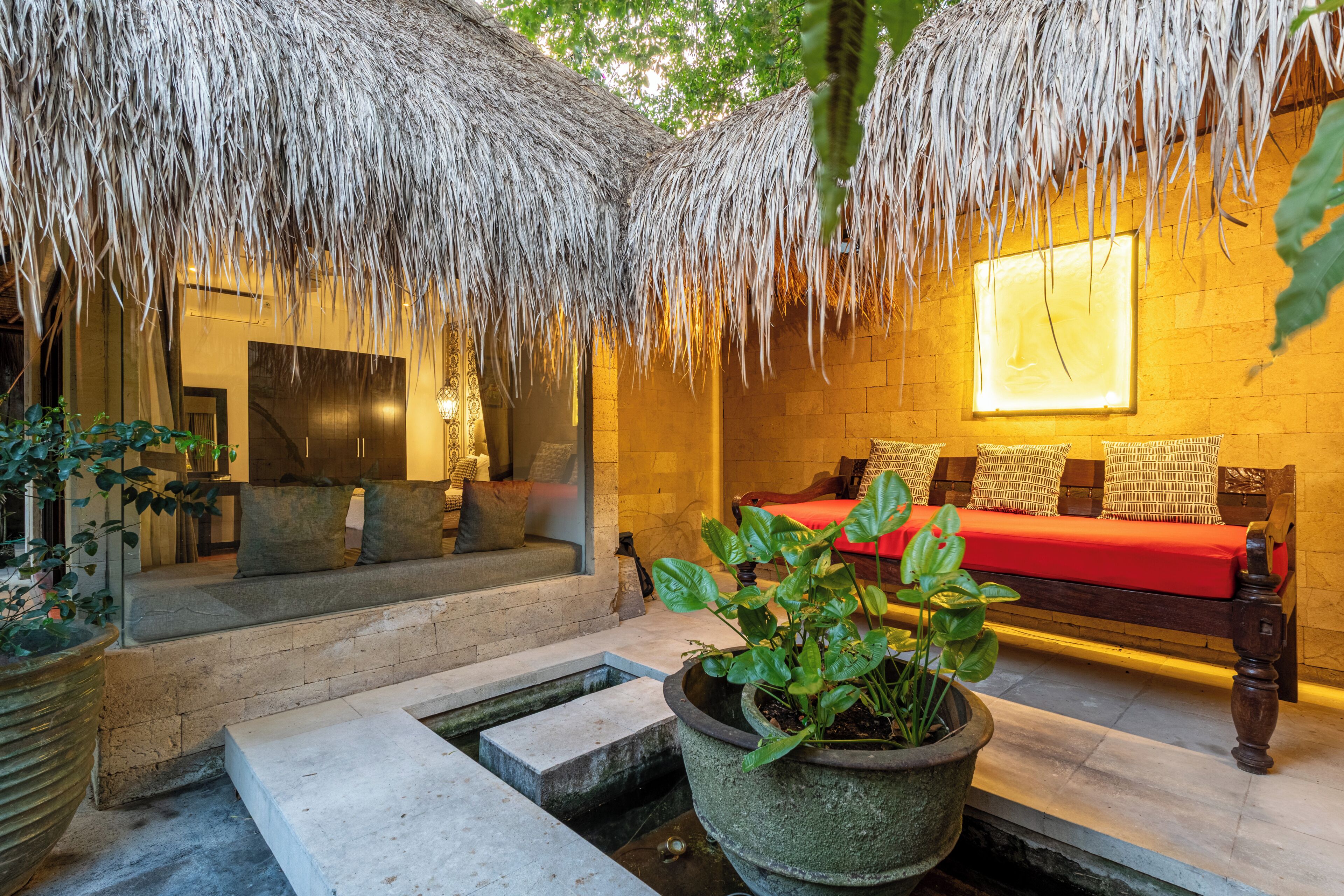 A room with a thatched roof, a bed with red bedding, a stone bench, and a potted plant.
