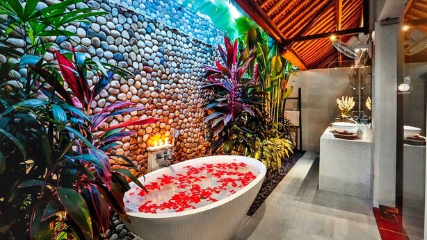Un moderno espacio de spa con una pared de piedra, una bañera grande llena de agua y pétalos de rosa, y varias plantas.