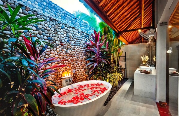 Un moderno espacio de spa con una pared de piedra, una bañera grande llena de agua y pétalos de rosa, y varias plantas.