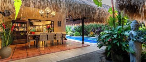 A restaurant with a thatched roof, indoor seating, and a pool area.
