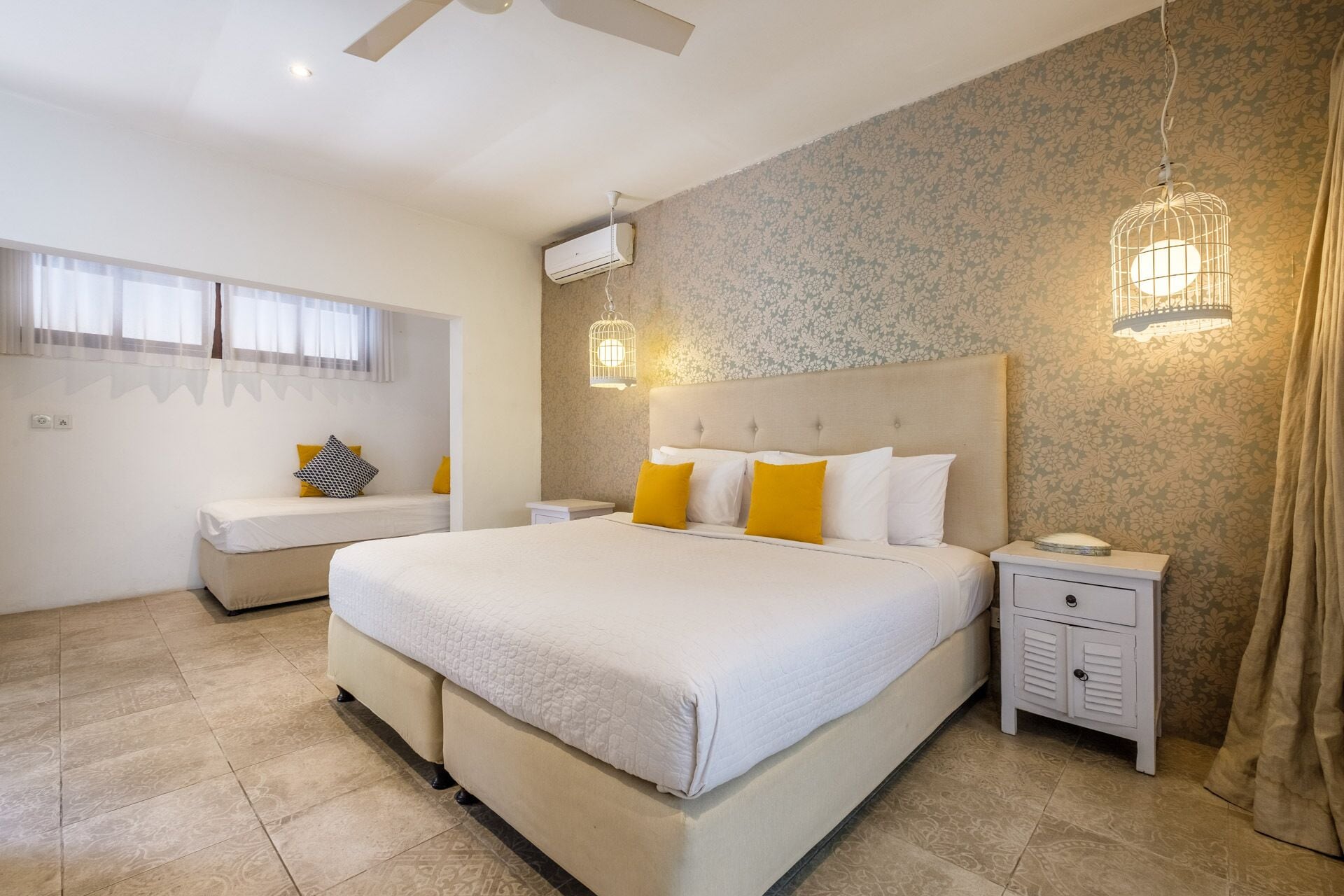 A bedroom with a large bed, two bedside tables, a nightstand, a ceiling fan, and a window with sheer curtains.