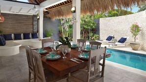 Comedor con una mesa de madera dispuesta para cuatro personas, rodeada de sillas de mimbre. Se ve una piscina al fondo, además de plantas y una pared blanca cerca.