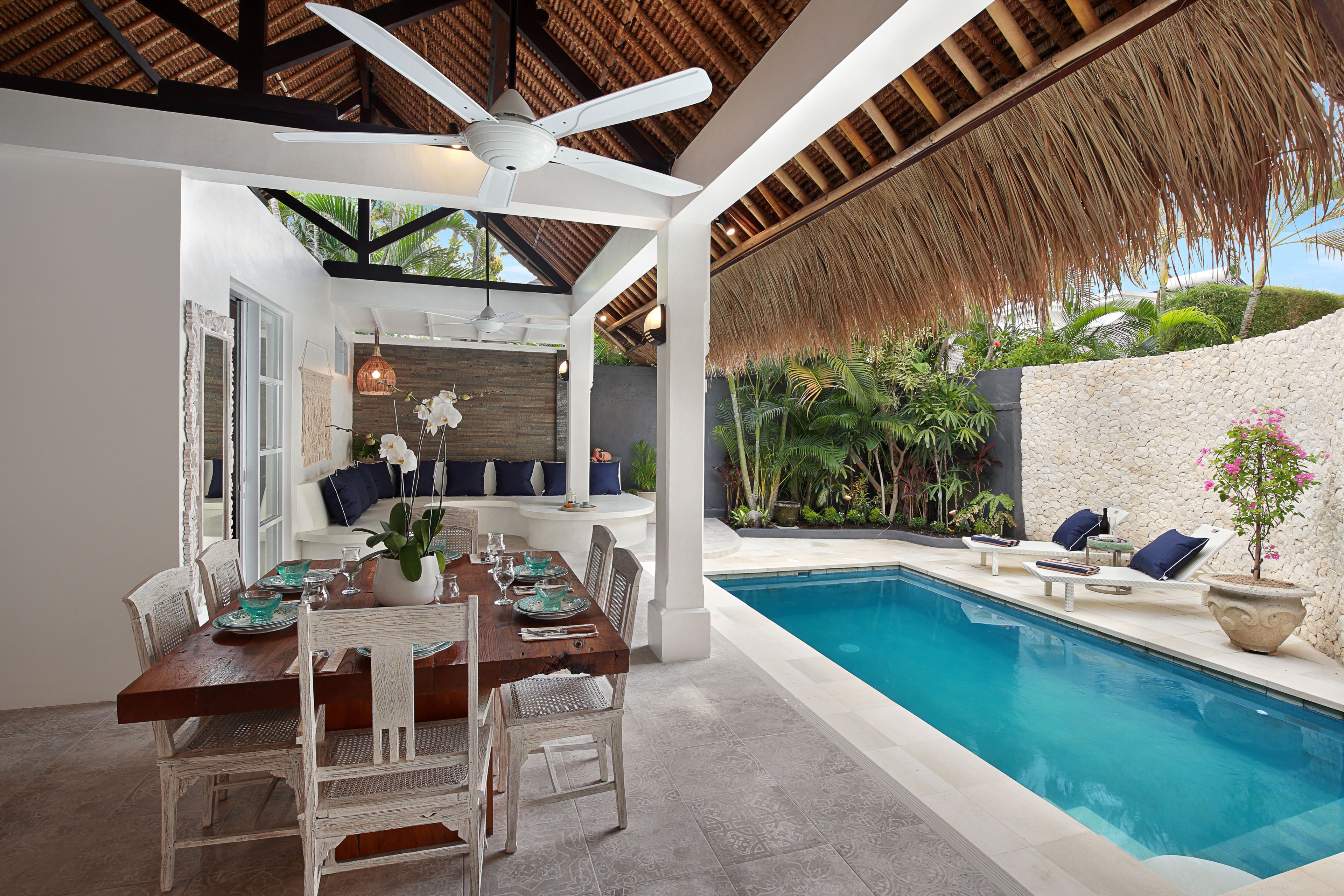 Un espace repas avec vue sur la piscine, comprenant une table en bois, des chaises blanches et un ventilateur de plafond.