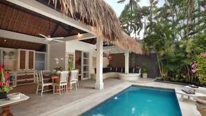 Un espace piscine doté d’un toit de chaume, un coin repas avec des chaises blanches et une vue sur les palmiers.