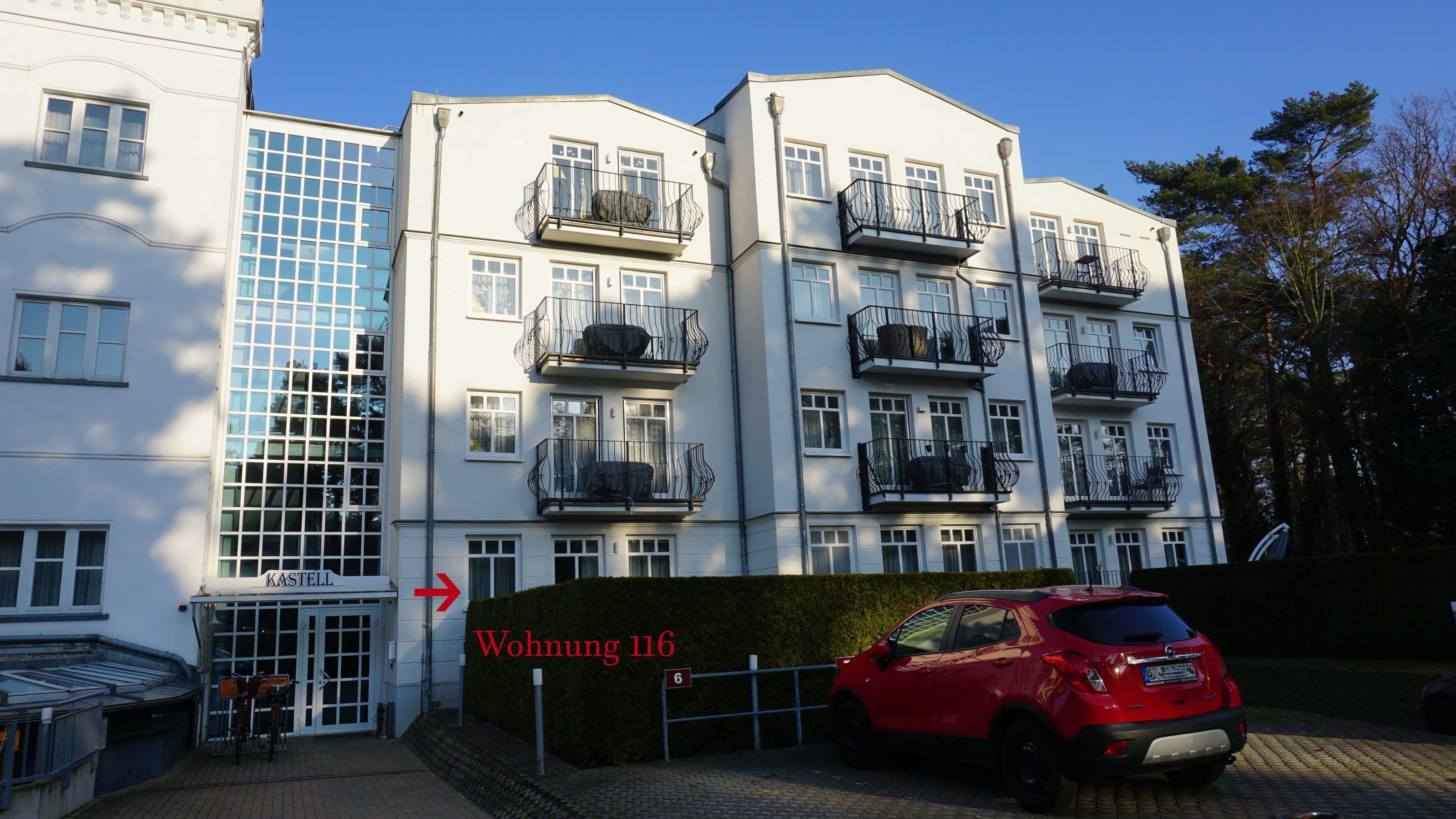 A multi-story building with a white facade, balconies with metal railings, and a red car parked in front.