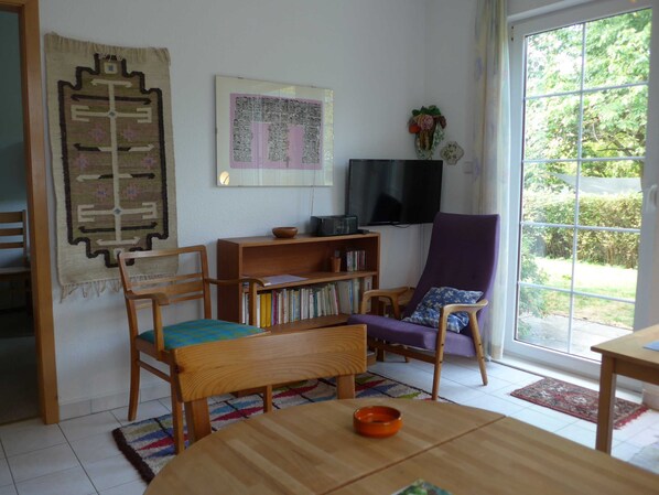 Un salon avec un canapé en bois, une chaise en bois, une petite table sur laquelle se trouve un bol, une bibliothèque, un téléviseur et une fenêtre donnant sur un paysage verdoyant.