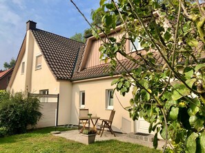 Une maison à deux étages avec un toit en tuiles, un petit patio avec une table et des chaises, et un jardin.