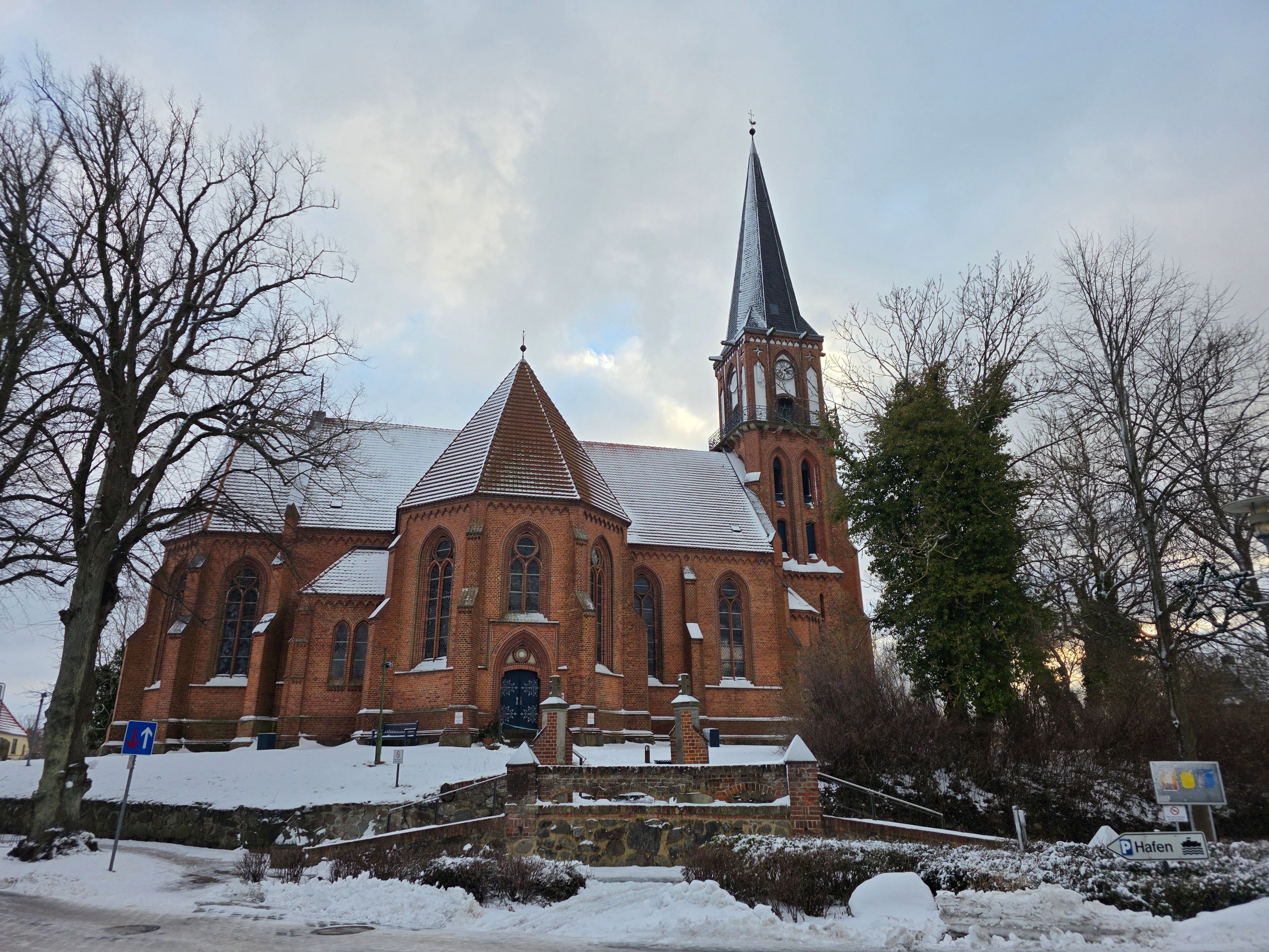 Une église dotée d’un clocher élancé et d’une tour horlogée, entourée d’arbres couverts de neige.