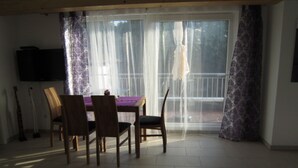 Ein Essbereich mit Tisch und Stühlen, einem Fernsehgerät an der Wand und Blick durch ein Fenster mit durchsichtigen Vorhängen.