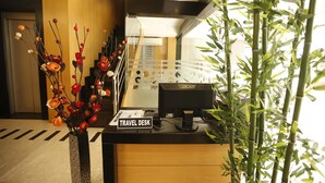 Un mostrador de recepción con un letrero de oficina de turismo, un jarrón decorativo con flores y una planta de bambú.