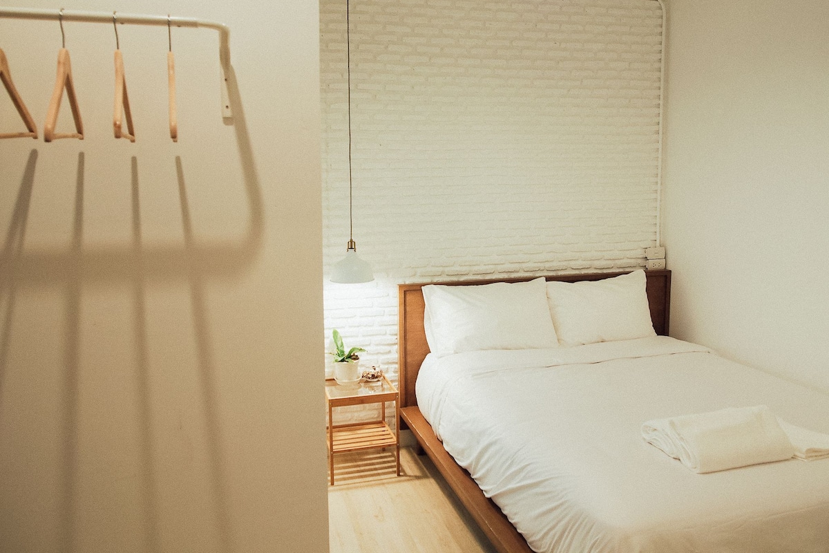 Quarto com cama de madeira, roupa de cama branca, luminária pendente e parede de tijolos.