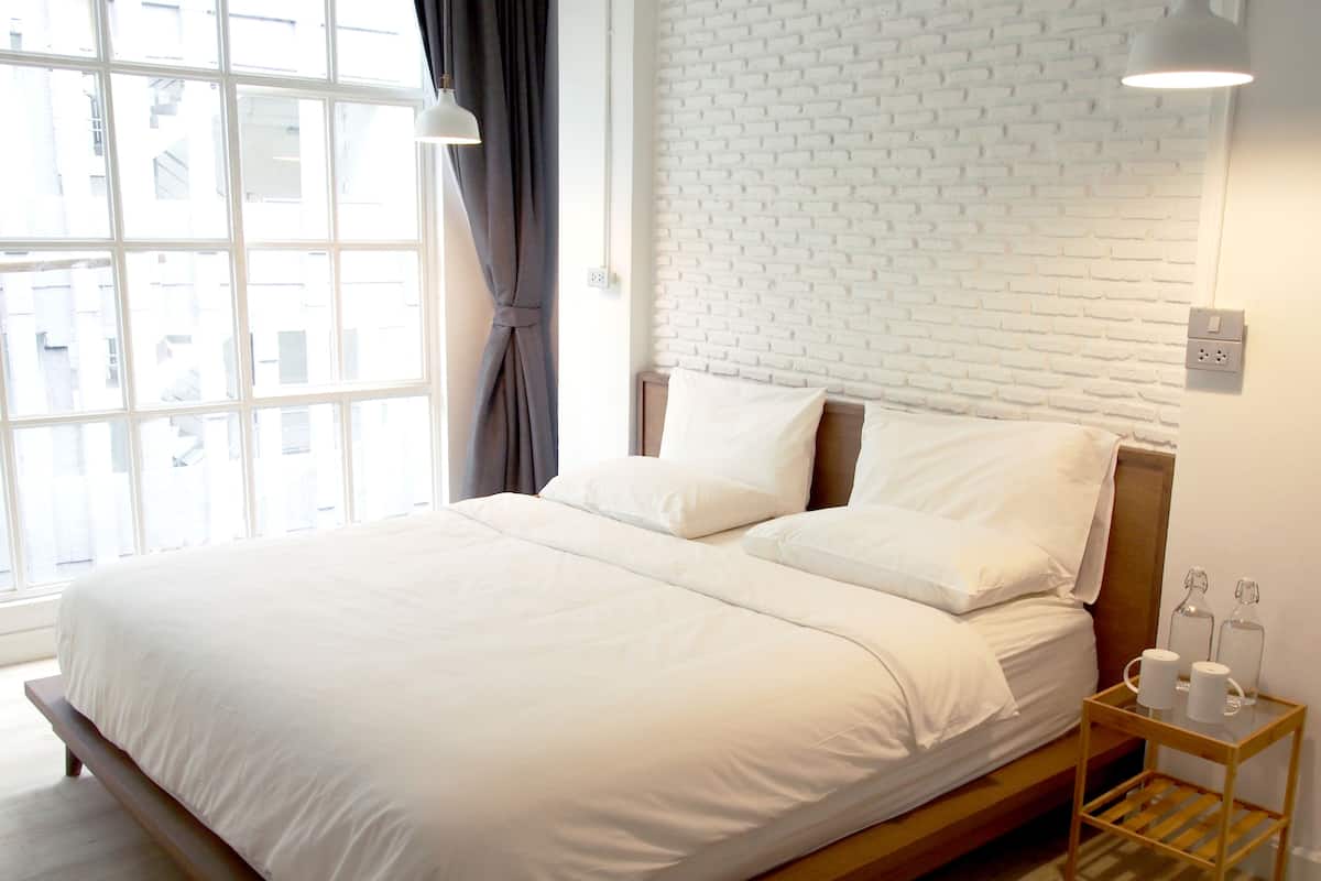 Cama bem arrumada com lençóis brancos, cabeceira de madeira e mesa de cabeceira com duas garrafas de vidro e uma caixa de lenços.