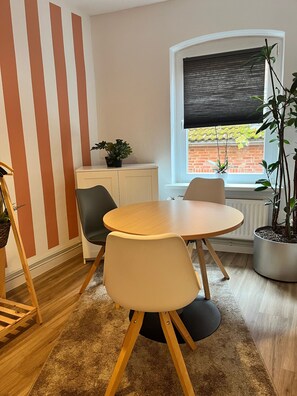 A dining area with a round table, two chairs, a cabinet, and a window with a view of a brick wall and plants outside.