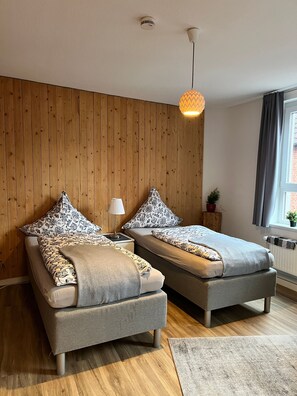 Un dormitorio moderno con dos camas, paneles de madera en la pared, una lámpara colgante y un ventanal con cortinas.