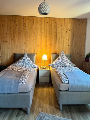 A room with two beds, a wooden wall, a hanging lamp, and a small table with a lamp.