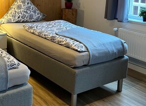 A neatly made bed with floral-patterned bedding, a grey upholstered bench, and a wooden bedside table with a red pot. - Apartment / apartment 2 - Buxtepartment (Buxtehude)