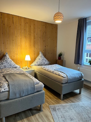 A bedroom with two beds, wooden panel walls, a hanging light fixture, a window with curtains, and a radiator.
