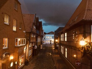 A narrow cobblestone street lined with traditional European buildings, some with illuminated signs, under a twilight sky.