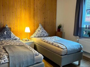 A room with two beds, a wooden wall, a window with curtains, and a small potted plant.