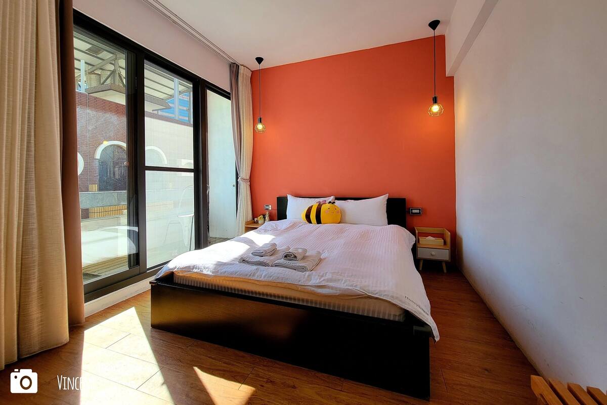 A bedroom with a bed, a nightstand, a lamp, and a view of the outside.