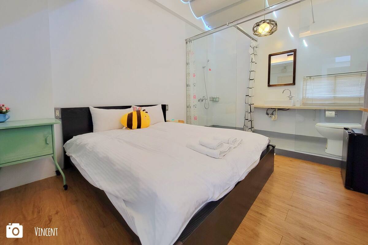 A modern bedroom with a large bed, a glass shower enclosure, a green nightstand, and a white bathroom with a sink and mirror.