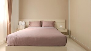 Una cama bien tendida, con un cabecero de color claro, una mesita de noche con lámpara y una pequeña cesta a su lado.