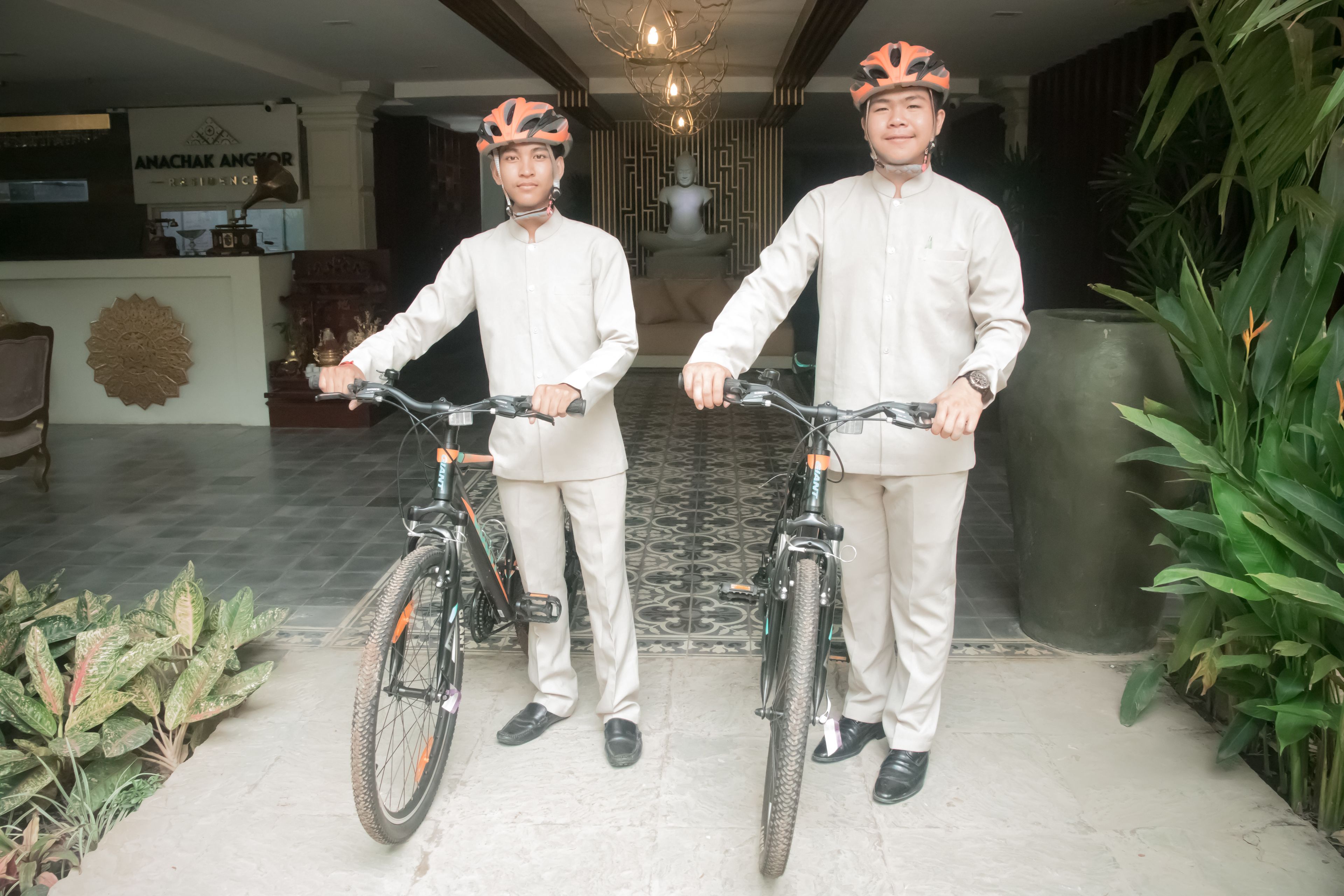 Two individuals in traditional attire holding bicycles in a lobby with a reception desk, decorative wall, and potted plants.