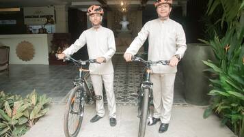 Two individuals in traditional attire holding bicycles in a lobby with a reception desk, decorative wall, and potted plants.