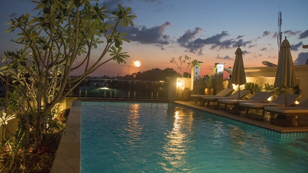 A swimming pool with lounge chairs and umbrellas, surrounded by greenery and a sunset in the background. - Anachak Angkor Suite (Siem Reap)