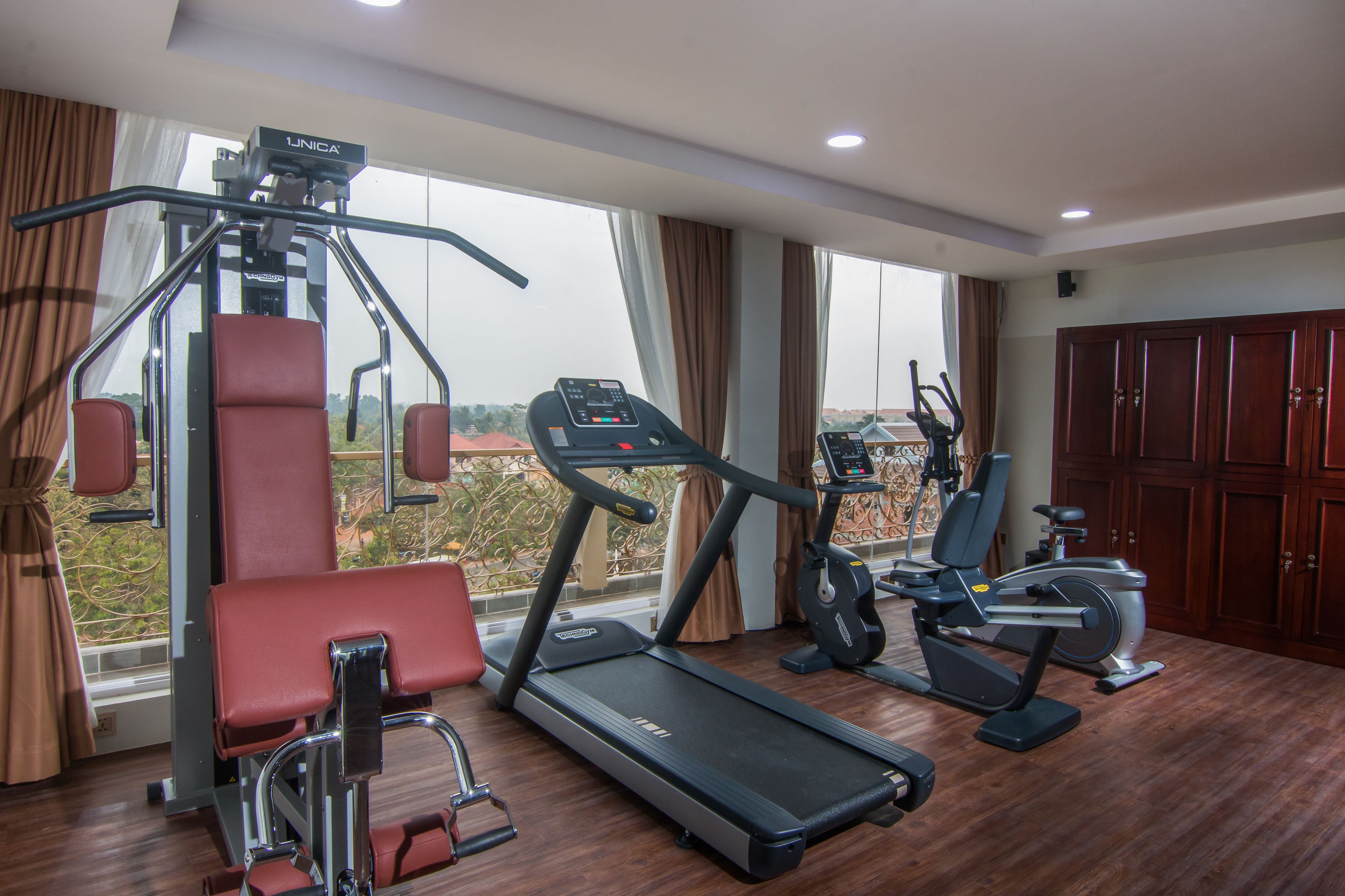 Un gimnasio con cinta de correr, elíptica y máquina de pesas, ventanas amplias con vistas y suelo de madera.
