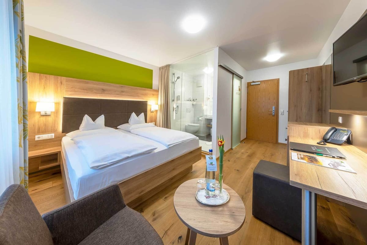 A hotel room with a bed, a desk with a telephone, a chair, and a small table with a bottle and glasses.