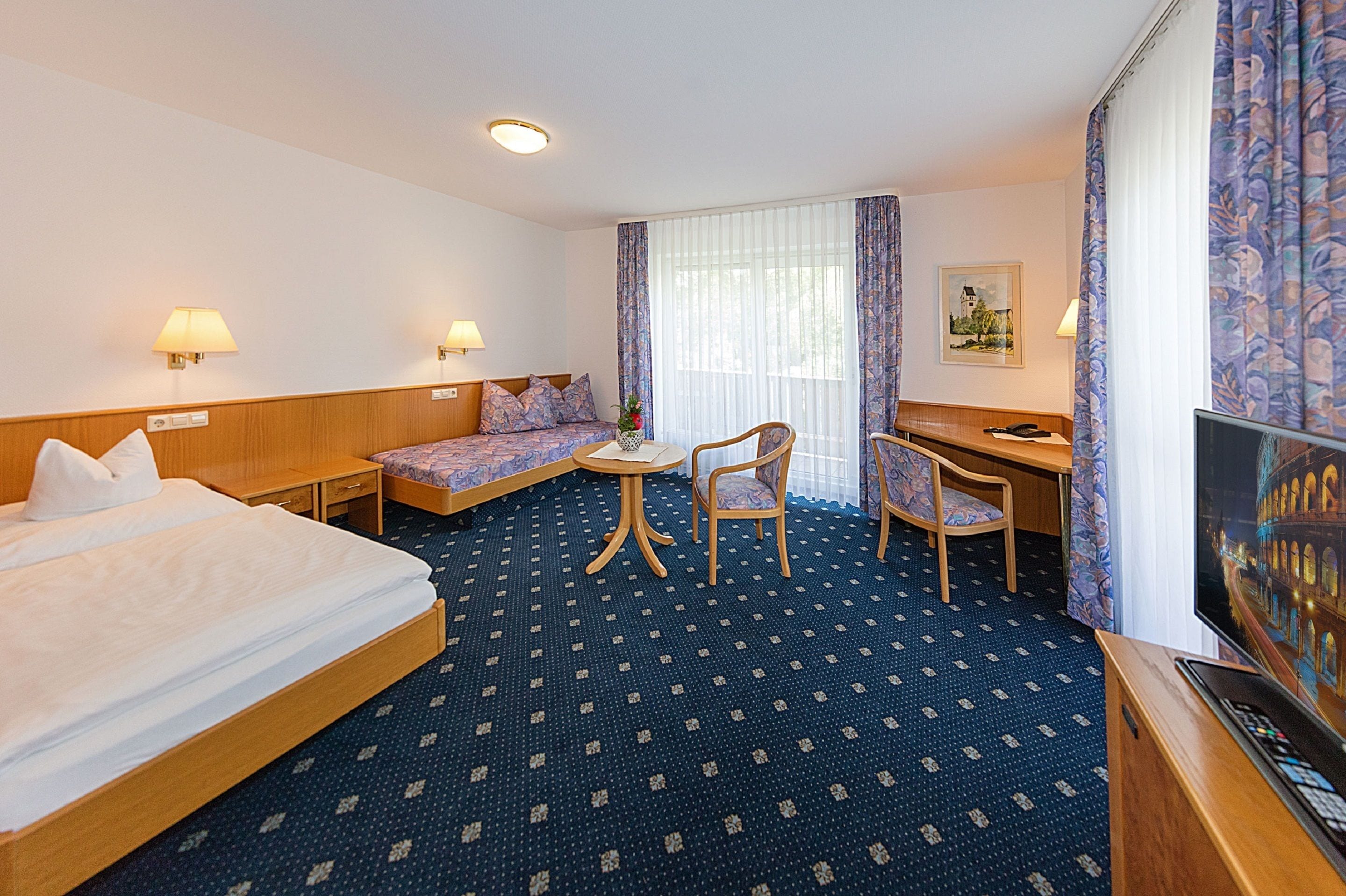 A hotel room with a bed, a desk, a chair, a small table, and a television.