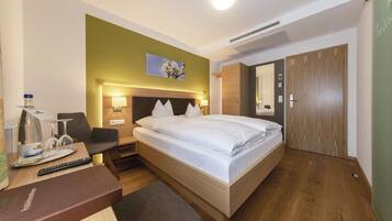 A hotel room with a bed, bedside table, chair, and a desk with a remote control.
