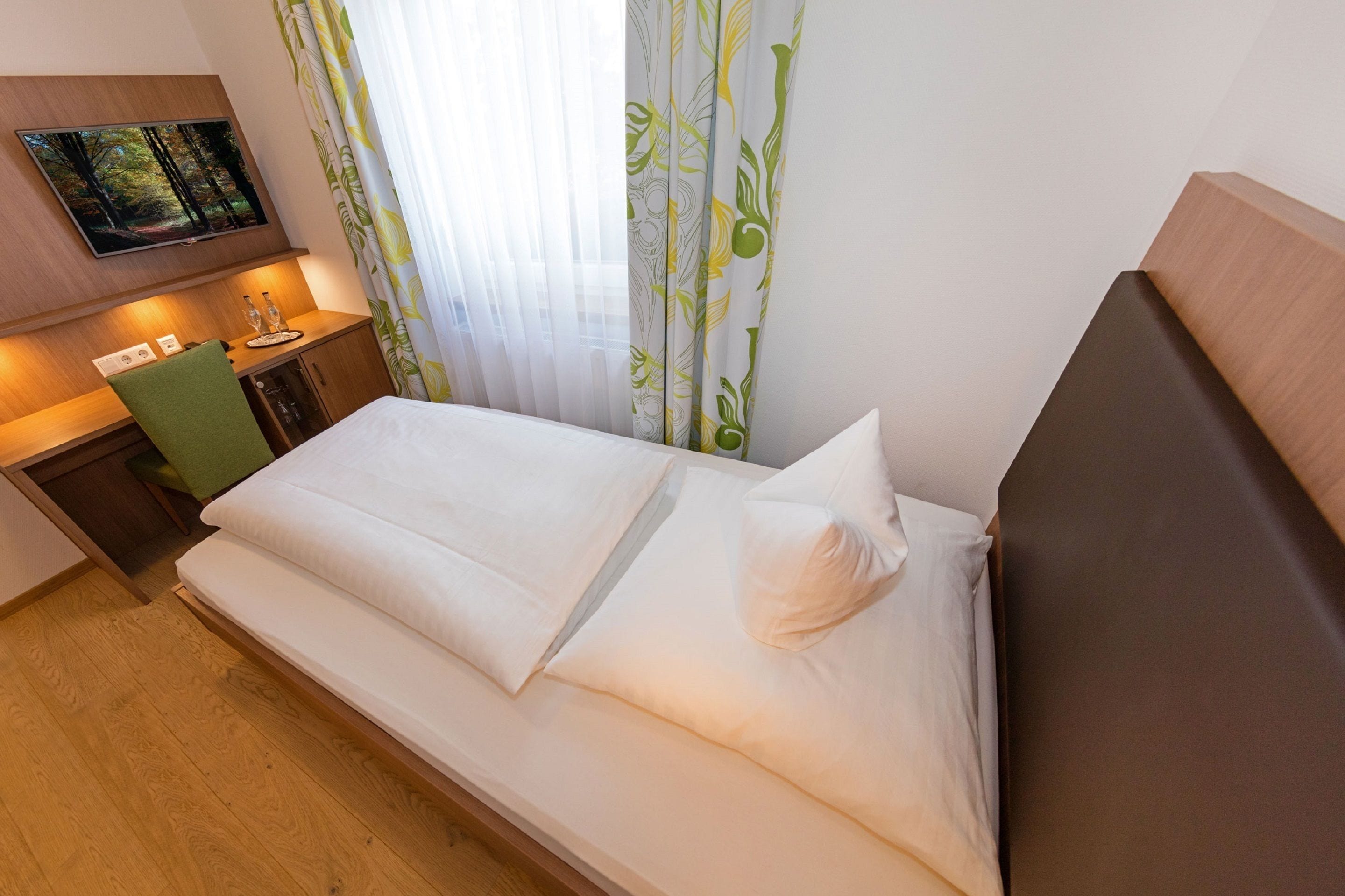 A hotel room with a bed, a desk, a chair, a TV mounted on the wall, and curtains with a leaf pattern.