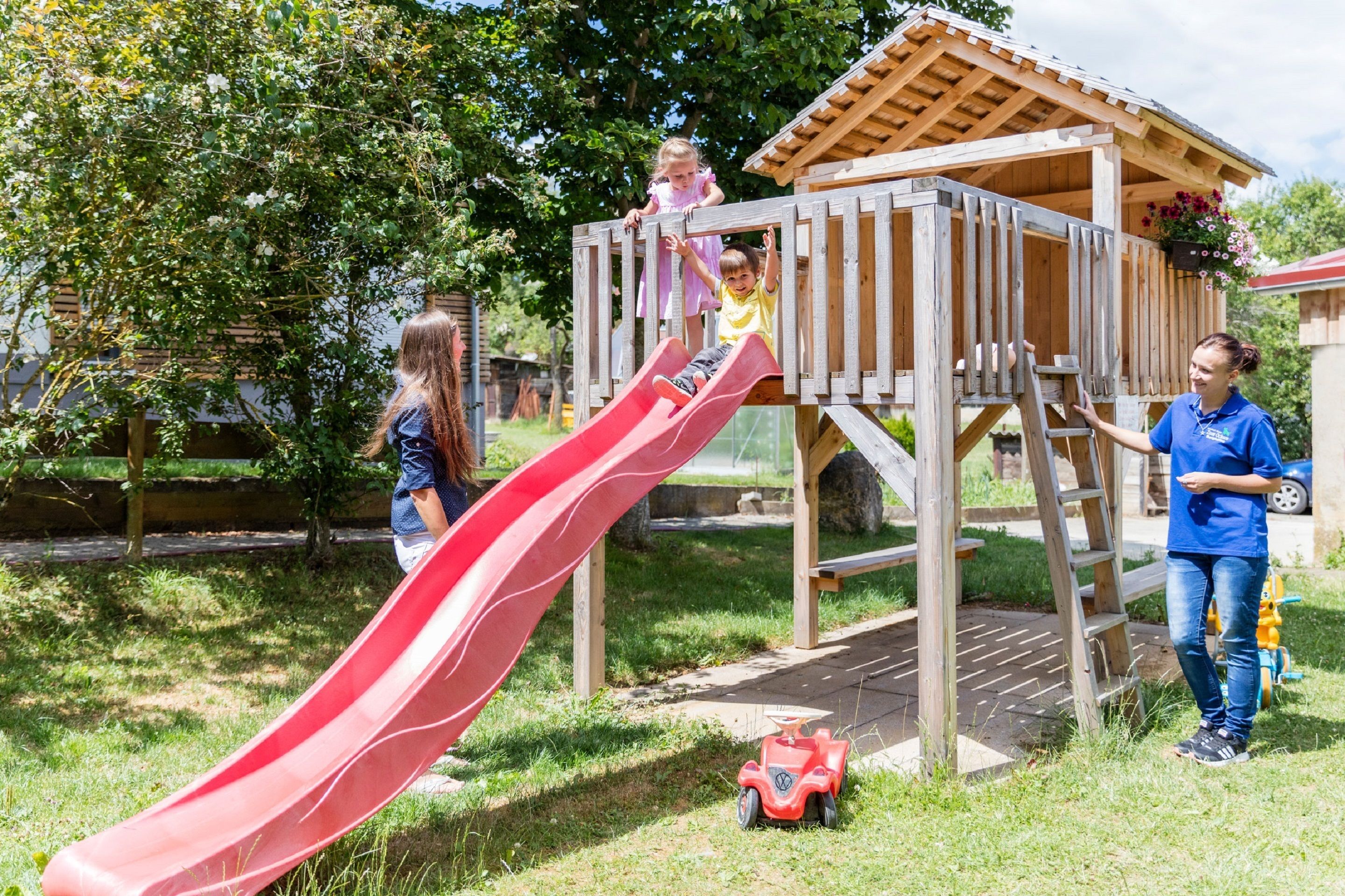 A wooden playhouse with a red slide, surrounded by greenery and a paved area.