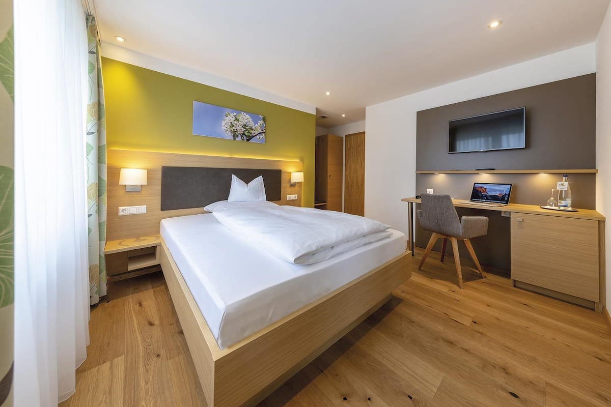 A modern hotel room with a large bed, a desk with a laptop, a flat-screen TV, and a wall-mounted artwork.