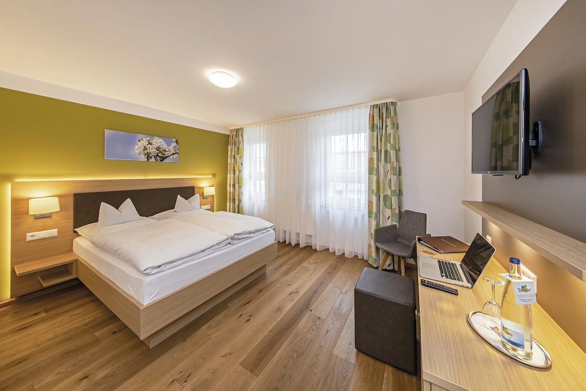 A hotel room with a bed, bedside tables, a desk with a laptop, a TV mounted on the wall, and a bottle of water on the desk.