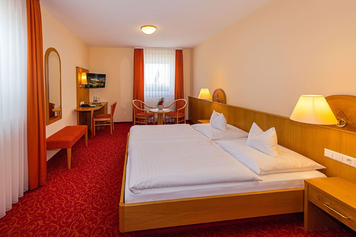 A hotel room with a large bed, a desk with a chair, a small table with chairs, a TV, and a window with curtains.