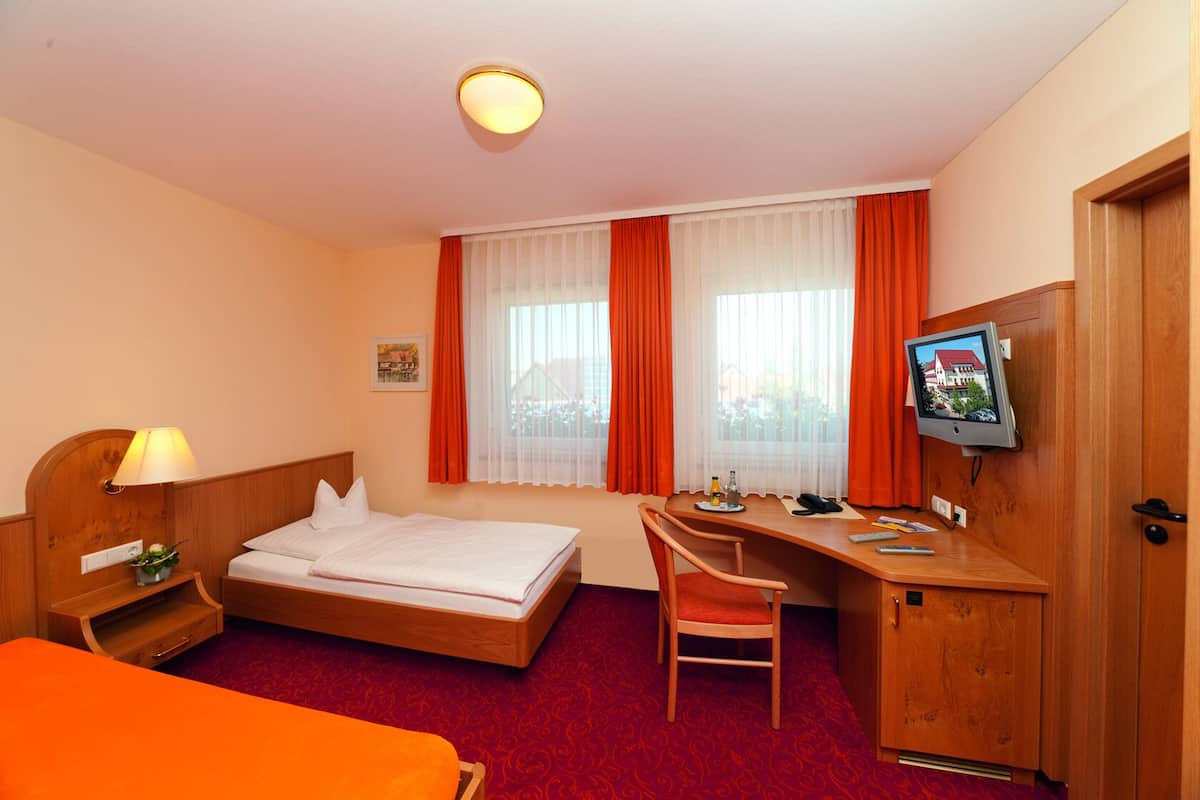 A hotel room with a bed, desk, chair, TV, and a small table with a lamp.