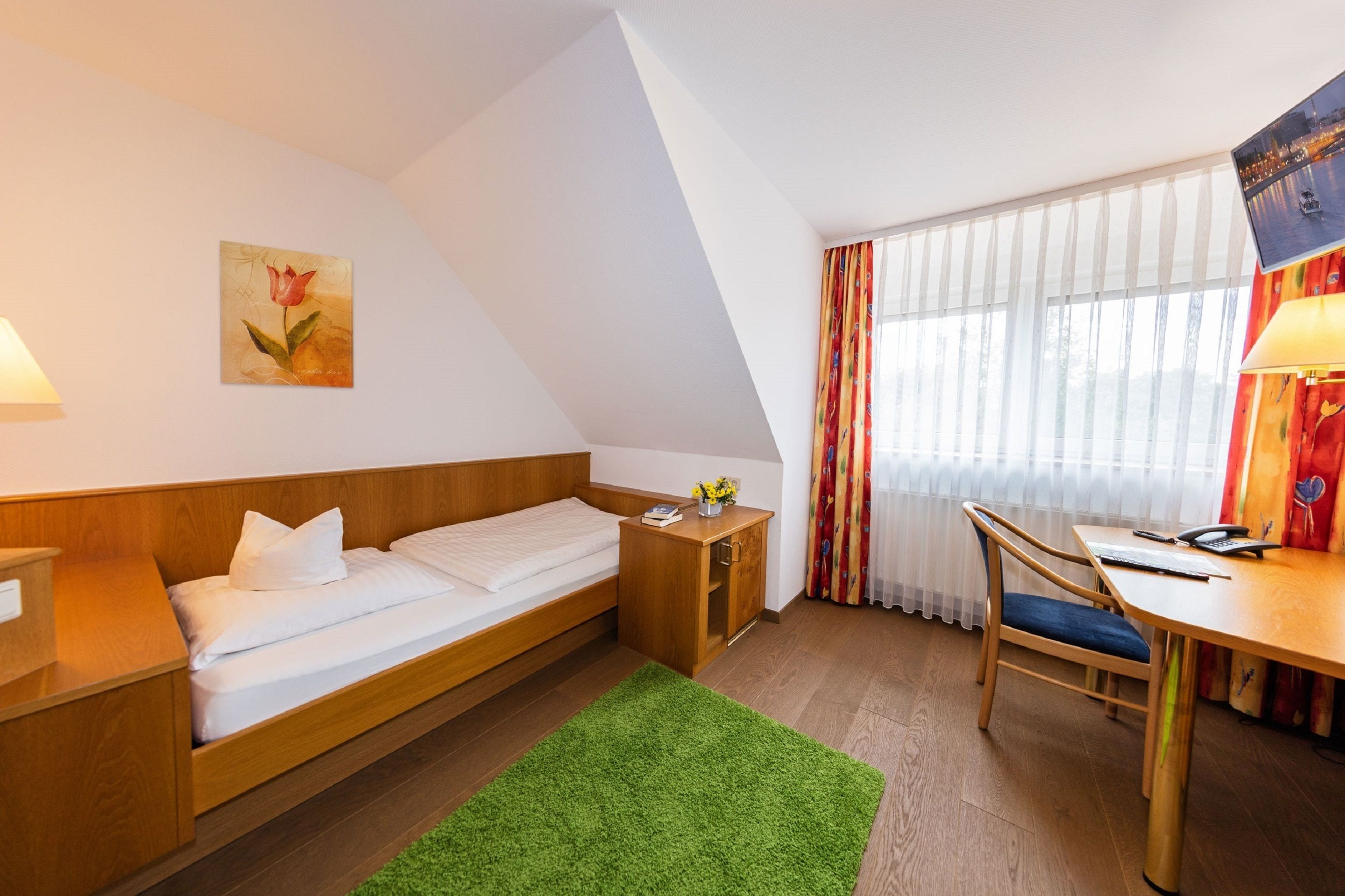 A modern hotel room with a bed, desk, chair, and a painting on the wall.