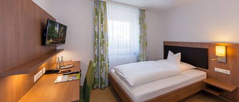A hotel room with a wooden desk, a flat-screen TV, a bed with white linens, a nightstand with a lamp, and a window with sheer curtains.