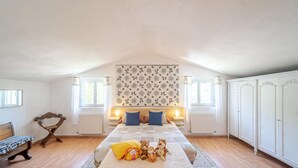 Une chambre spacieuse située dans les combles, avec un plafond haut, un grand lit, une armoire et une fenêtre avec des rideaux.