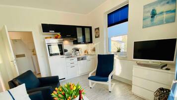Ein modernes Wohnzimmer mit einer Couch, einem Couchtisch mit Blumen und einer Flasche, einem Fernseher und einer Küche im Hintergrund.