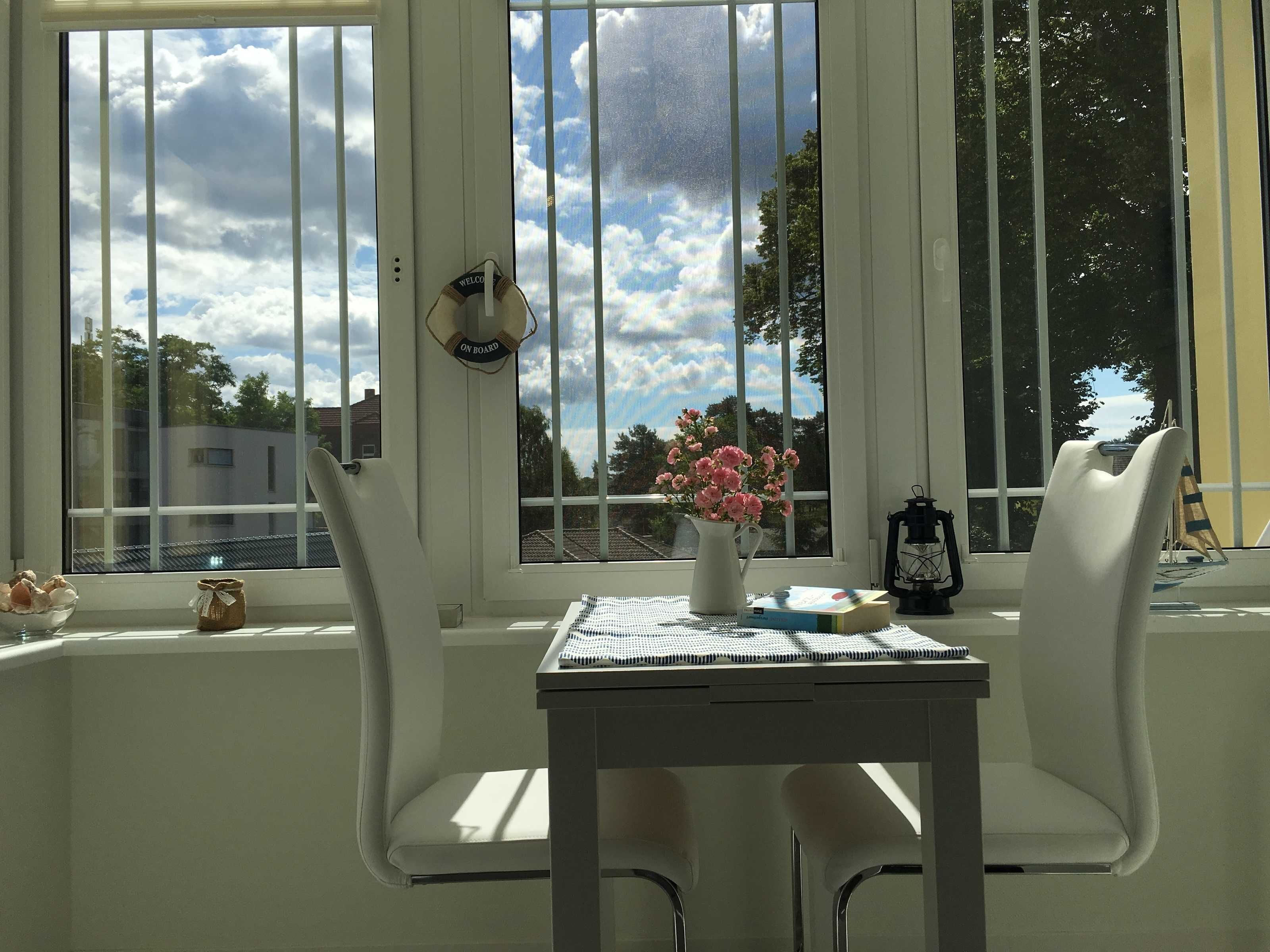 Ein Zimmer mit Tisch und zwei Stühlen, einem Fenster mit Blick auf Bäume und Gebäude, einer Vase mit Blumen und einer Laterne.