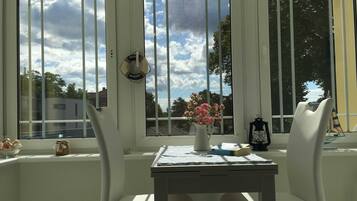 Ein Zimmer mit Tisch und zwei Stühlen, einem Fenster mit Blick auf Bäume und Gebäude, einer Vase mit Blumen und einer Laterne.