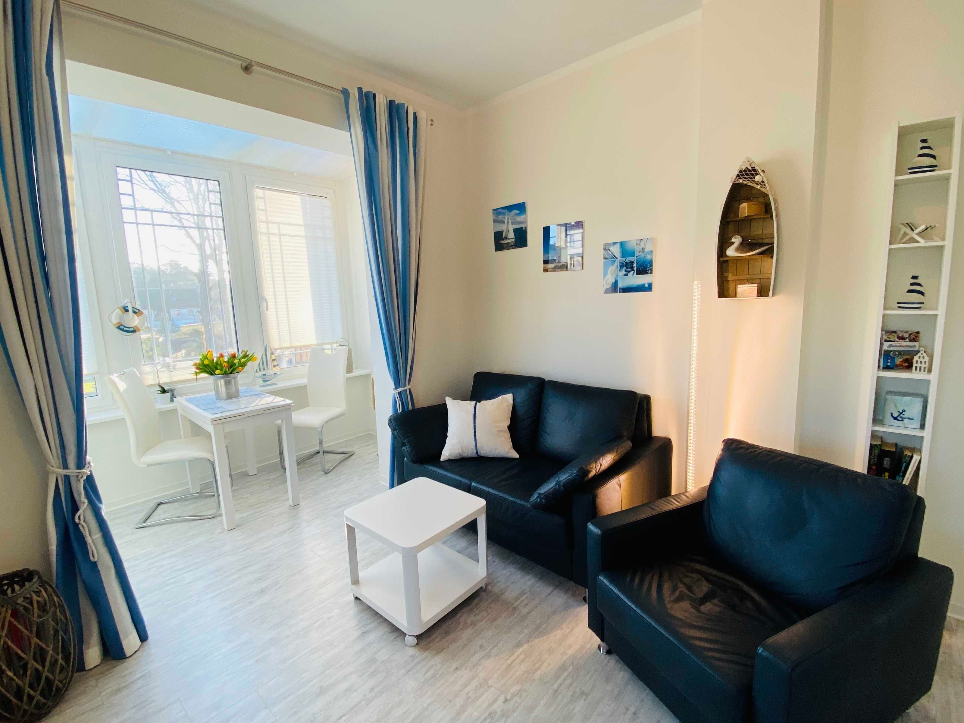 Ein Wohnzimmer mit einer schwarzen Ledersofa, einem weißen Couchtisch, einem Essbereich mit weißem Tisch und Stühlen sowie einem Bücherregal mit maritimer Dekoration.