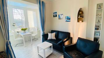 Ein Wohnzimmer mit einer schwarzen Ledersofa, einem weißen Couchtisch, einem Essbereich mit weißem Tisch und Stühlen sowie einem Bücherregal mit maritimer Dekoration.