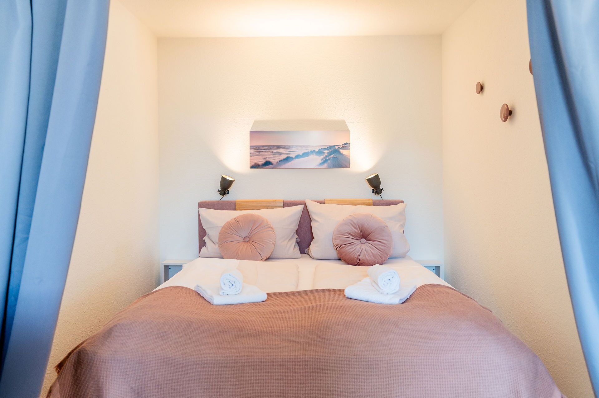 Ein ordentlich bezogenes Bett in Rosa und Weiß, ein Wandbild und ein kleiner Nachttisch mit Lampe.